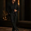 Full-length male model wearing a tailored black suit, black shirt, and polished dress shoes, posed in an elegant dark luxury interior with warm ambient lighting, arched walls, a patterned rug, and premium furnishings.