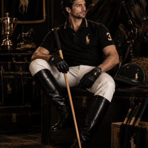 Close-up portrait of a man wearing a premium black polo shirt with subtle gold details, holding a polo mallet, with a horse and luxury equestrian setting in the dark background.