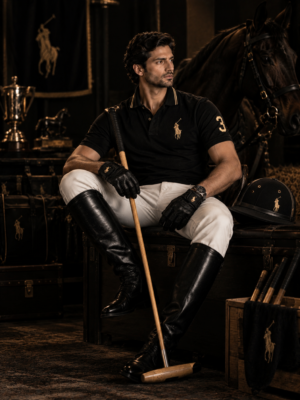 Close-up portrait of a man wearing a premium black polo shirt with subtle gold details, holding a polo mallet, with a horse and luxury equestrian setting in the dark background.